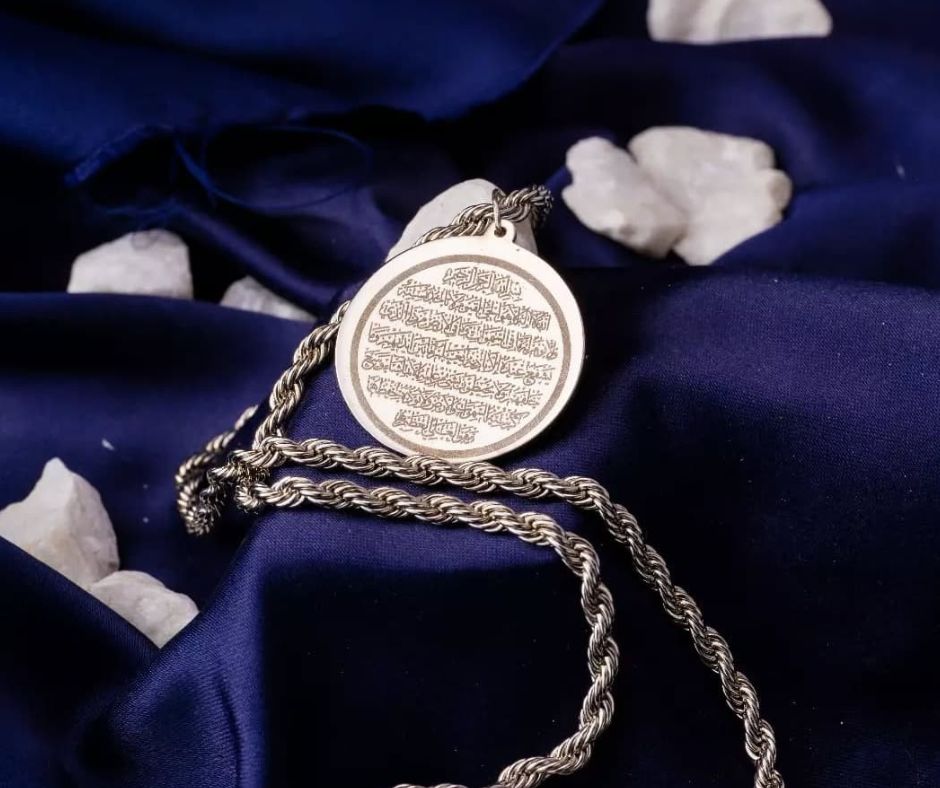 Silver Ayat ul Kursi pendant with intricate calligraphy on a twisted chain against dark blue fabric