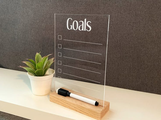Clear acrylic to do list board with wooden base and marker next to small potted plant on white shelf