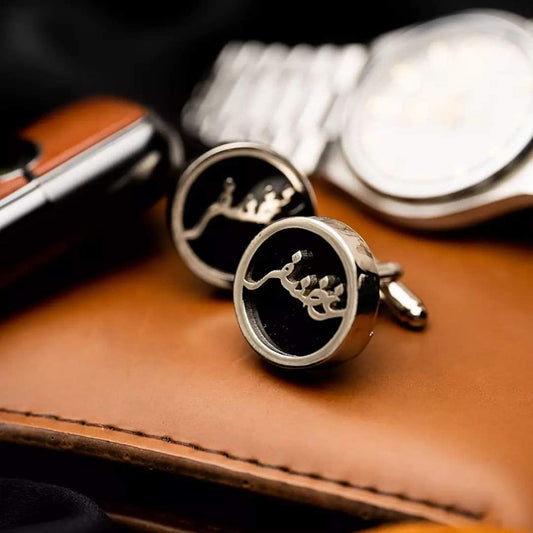 Silver round custom name cufflinks with black background placed on brown leather surface beside watch and car key